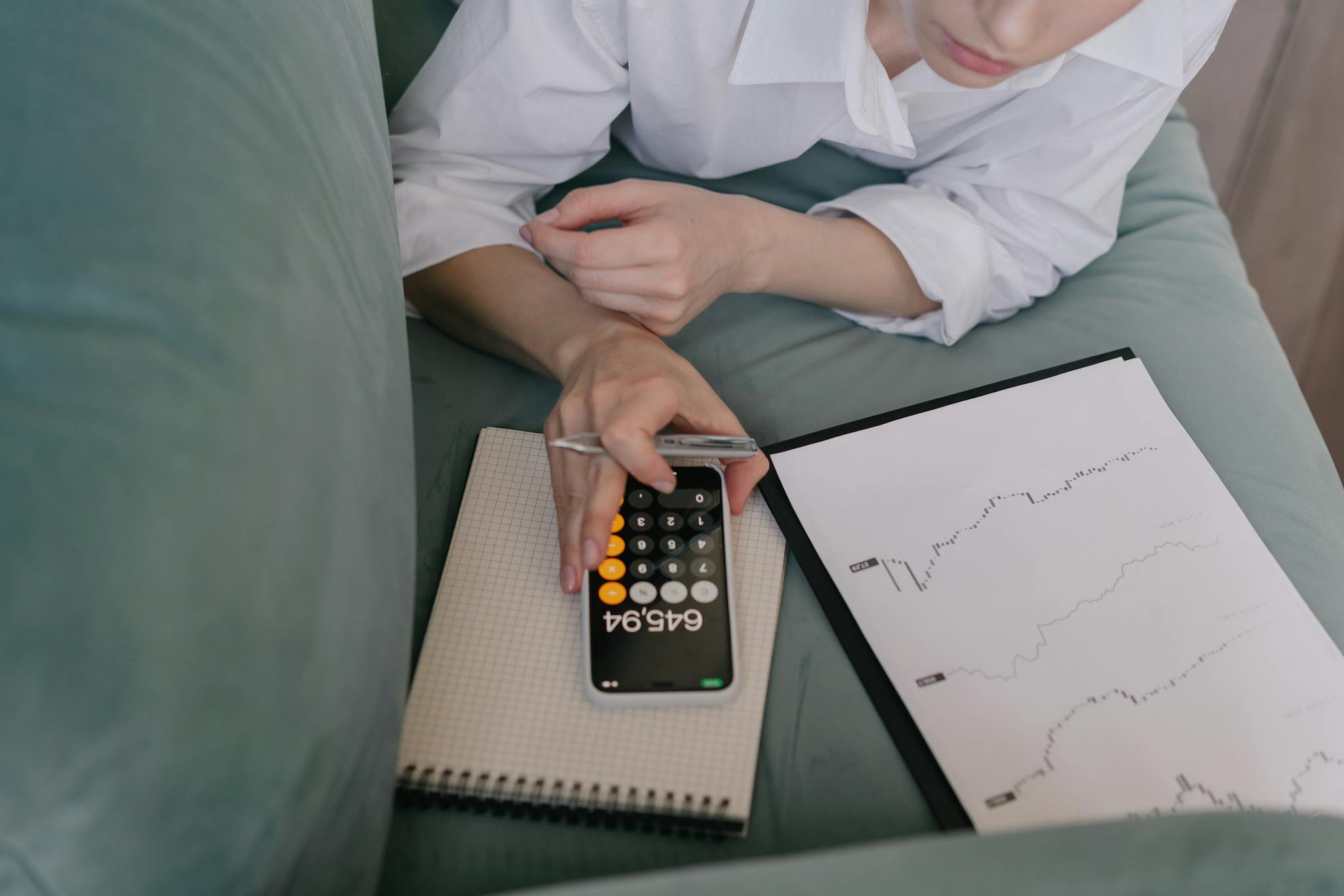 A woman works indoors using a smartphone calculator and analyzing financial graphs, symbolizing modern finance.